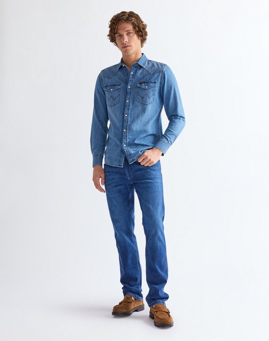 Man met krullend haar in een blauw denim overhemd en Wrangler jeans, staand op witte achtergrond.