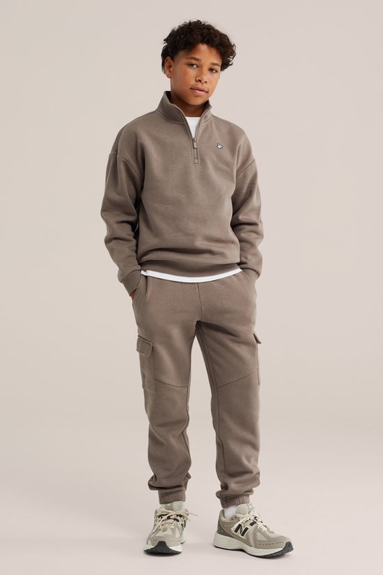 Jongens outfit: bruine trui met halve rits en logo, bruine joggingbroek met cargo zakken, witte t-shirt en grijze schoenen.