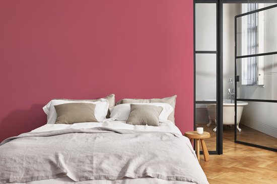 Een slaapkamer met een roze muur, een tweepersoonsbed met linnen beddengoed en kussens, en een glazen deur naar de badkamer.