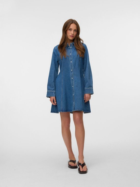 Model staat frontaal, draagt een blauwe denim jurk met lange mouwen en een uitlopende rok.