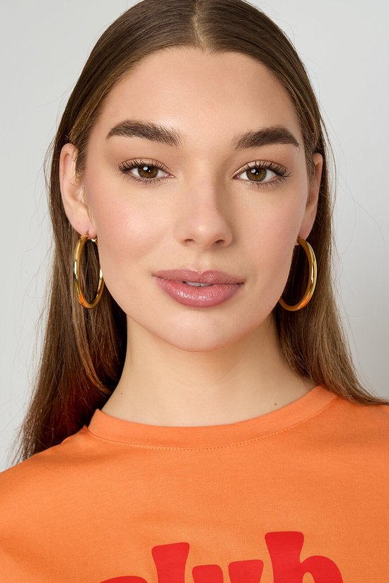 Model met gouden oorringen en oranje shirt.