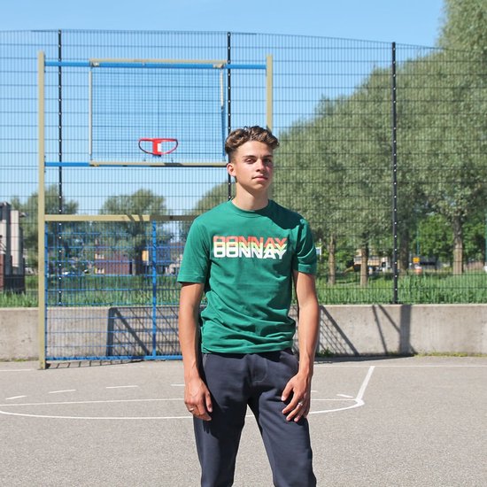 Jonge man staat op basketbalveld, draagt groene t-shirt met tekst en donkere joggingbroek.