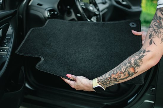Vrouw met tatoeages op arm houdt een zwarte vloermat voor een autodeur.