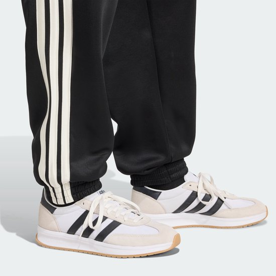 Detail van de broekspijpen van een zwarte trainingsbroek met witte strepen, gedragen met witte sneakers.