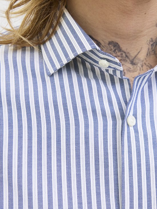 Een man met tatoeages op de nek, draagt een blauw-wit gestreept overhemd met witte knopen.
