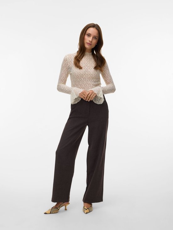 Model in een witte kanten top met lange mouwen en een hoge kraag, gecombineerd met een donkerbruine broek met wijde pijpen.