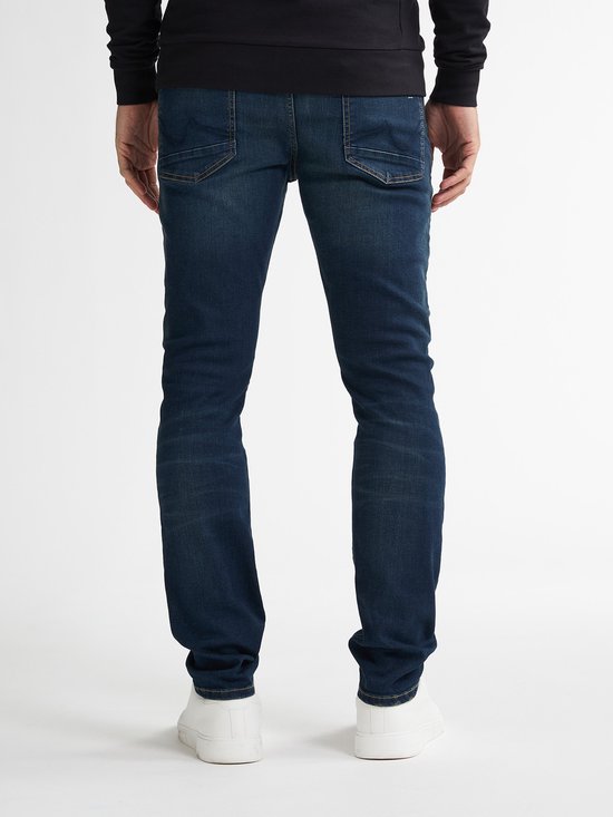 Man in donkerblauwe slim fit jeans met witte sneakers, van achteren gezien.