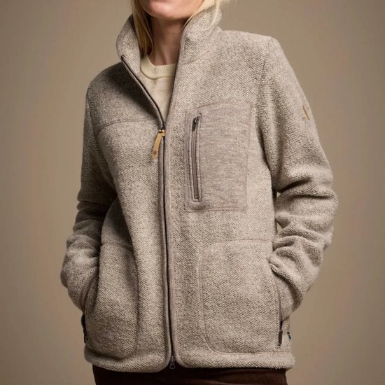 Model draagt een beige wollen vest met ritssluiting, opstaande kraag, borstzak en steekzakken.