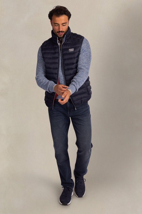 Man in donkerblauwe gewatteerde bodywarmer, blauw gebreid vest en jeans. Op de borst een Patch 'New Zealand Auckland'.