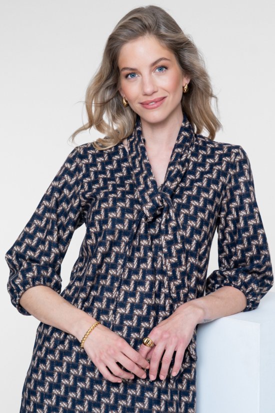 Close-up van een vrouw met blonde haren, draagt een navy jurk met geometrische print en gouden accessoires.