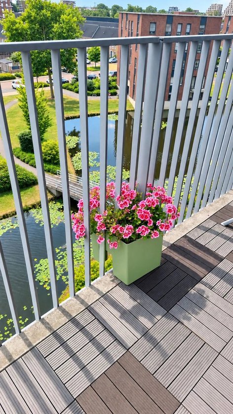 Olijfgroene plantenbak met felroze bloemen, geplaatst op een balkon met grijze en bruine tegels.