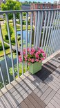 Olijfgroene plantenbak met felroze bloemen, geplaatst op een balkon met grijze en bruine tegels.