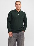 Man draagt donkergroen poloshirt met lange mouwen, kraag met knoopjes en geborduurd logo, met grijze jeans.
