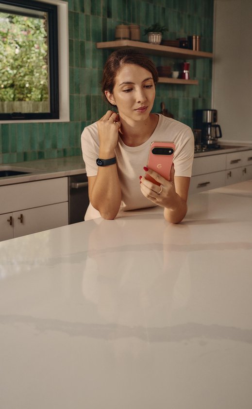 Vrouw met Fitbit Edge om de pols kijkt op haar smartphone in een moderne keuken.