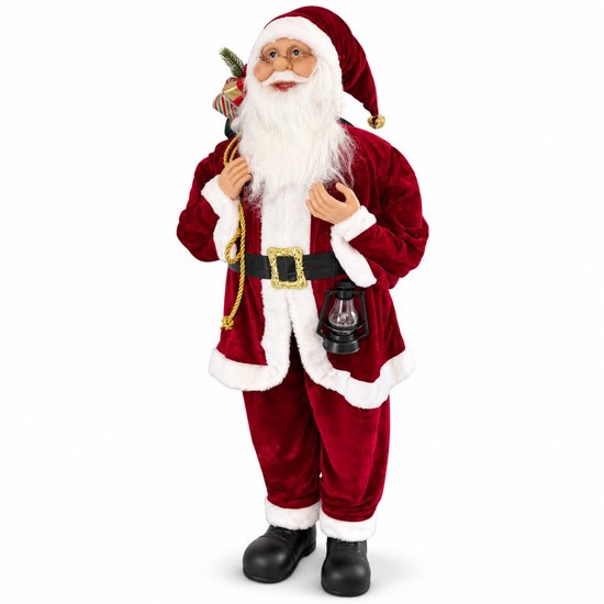 Père Noël debout en costume de velours rouge, avec barbe blanche, lunettes, lanterne et sac de cadeaux.