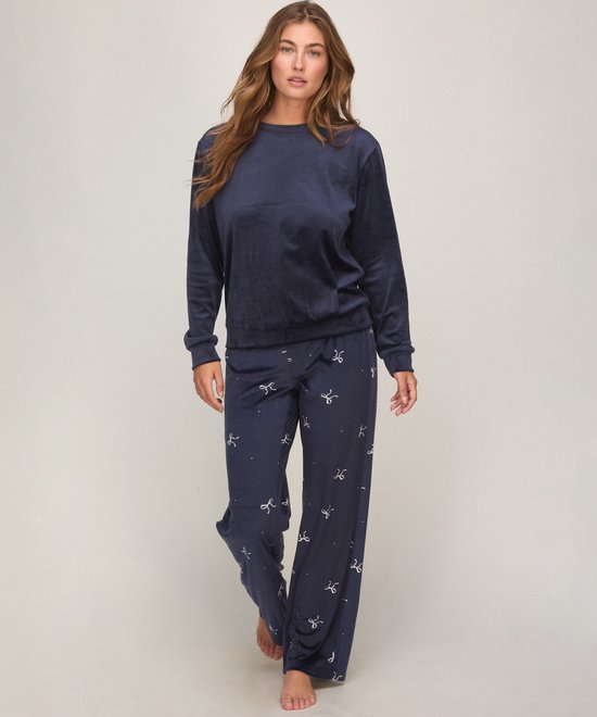Model draagt een marineblauwe velours pyjamaset met een effen trui en een broek met een wit strikjespatroon.