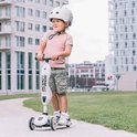 Kind met witte helm op een beige Scoot and Ride Highwaykick step in een stedelijke buitenomgeving.