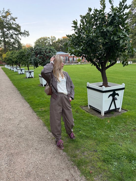 Vrouw draagt een taupekleurig Lofty Manner bomberjack, witte kanten top, wijde broek, schoudertas en sneakers in tuin.