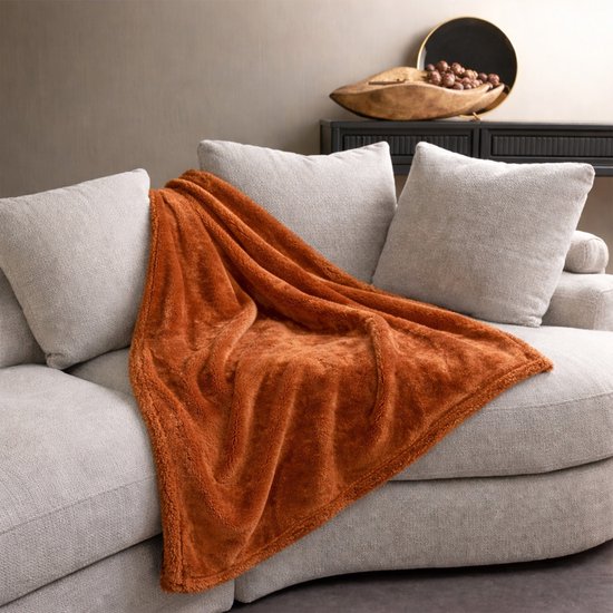 Een oranje terra plaid van fleece met teddy rand op een grijze bank.