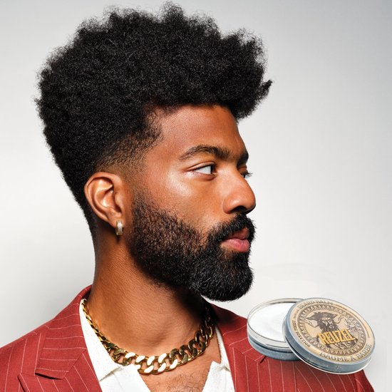 Man met donkere huid, volle baard en gouden ketting, naast een blikje Reuzel baardbalsem.