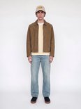Model volledig in beeld met bruin suède jack, beige t-shirt, blauwe spijkerbroek en pet.