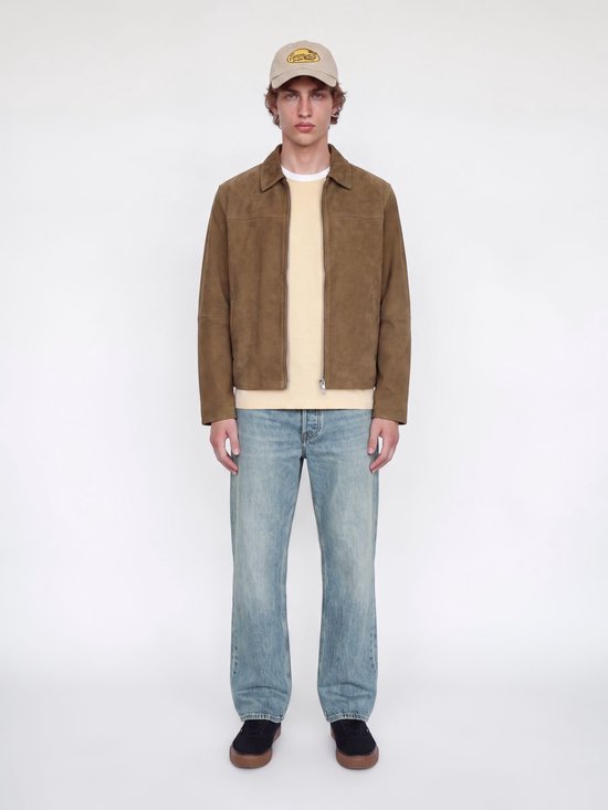Model volledig in beeld met bruin suède jack, beige t-shirt, blauwe spijkerbroek en pet.