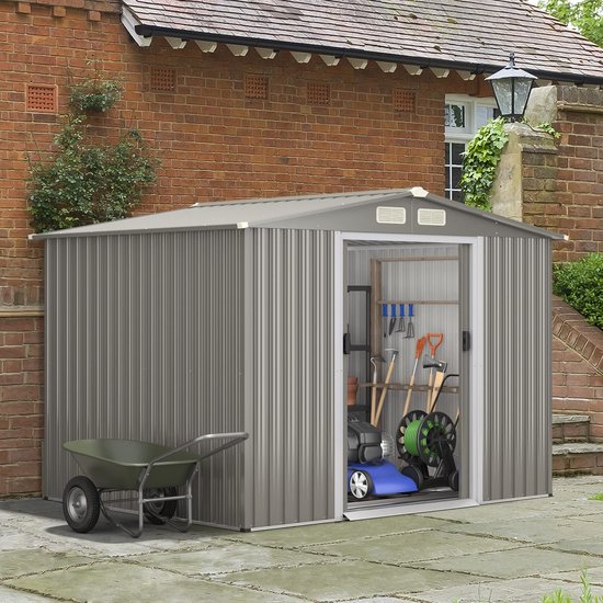 Grijze metalen tuinschuur op een terras voor een bakstenen muur, gevuld met tuingereedschap en een kruiwagen.