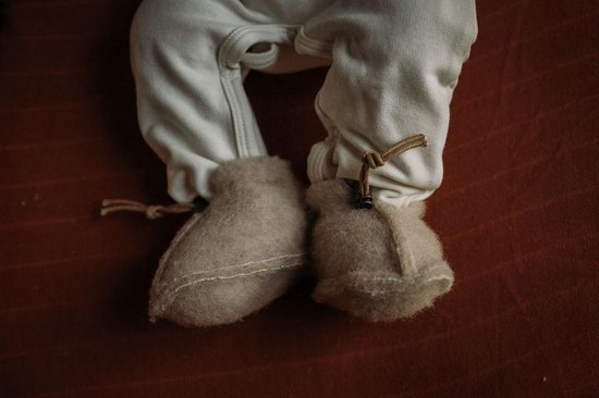Een baby draagt beige, wollen sloffen met bruine veterdetails. De sloffen zijn zacht en wollig.