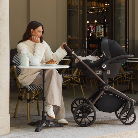 Vrouw zit aan tafel met een zwarte kinderwagen en baby-autostoel ernaast.