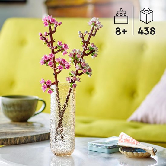 Twee takken met roze en witte kersenbloesems in een glazen vaas, met een mok en een ventilator op de achtergrond.