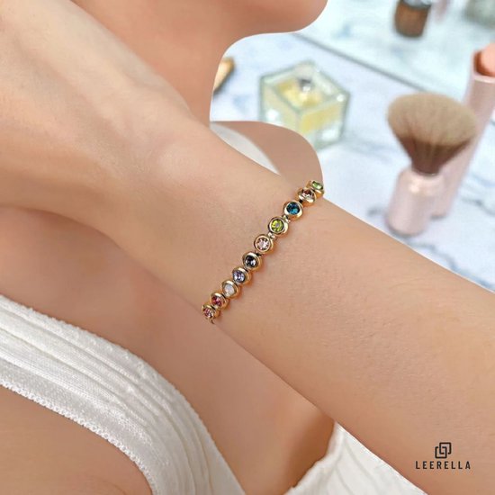 Arm van een vrouw met een goudkleurige armband met veelkleurige stenen, met wazige make-up spullen op de achtergrond.