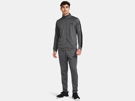 Man met grijze trainingsset: jack met zwarte strepen en logo, en broek met zwarte strepen en logo.