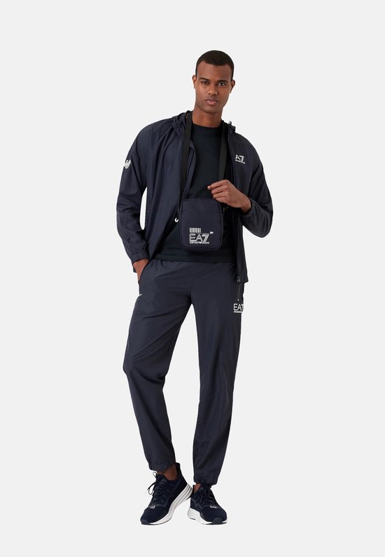 Een man draagt een marineblauw EA7 trainingspak met capuchon en een kleine schoudertas met EA7 logo.