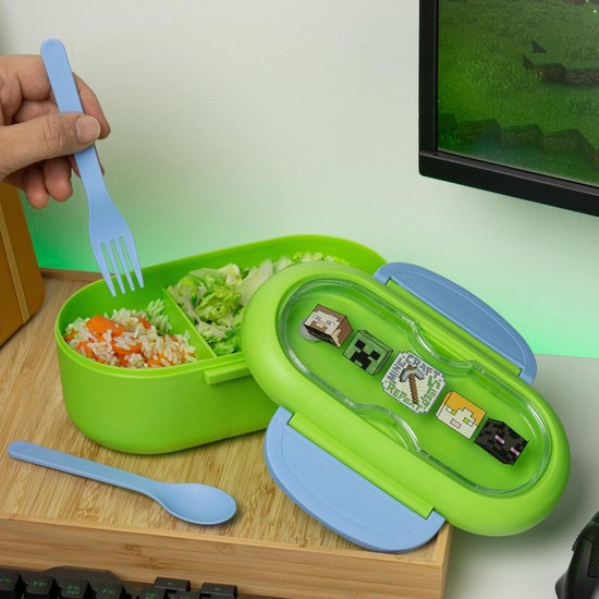 Groene lunchtrommel met Minecraft-personages, gevuld met rijst en salade. Blauwe vork in hand.