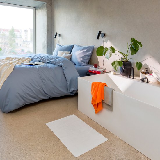 Lichtblauwe dekbedovertrek op een bed, met een gebreide beige plaid eroverheen gedrapeerd. Een oranje en grijze handdoek hangen over de rand van een wit bad.