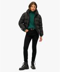 Vrouw in zwarte Superdry pufferjas met capuchon, groene coltrui, zwarte jeans en laarzen.