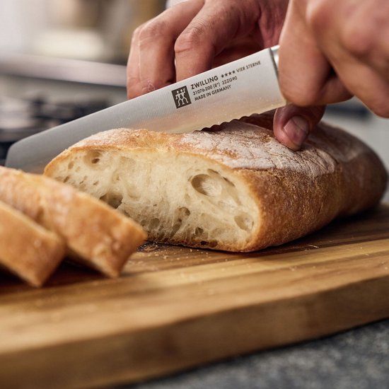 Handen snijden een brood met een kartelmes, met een zwart handvat. Er liggen gesneden stukken brood op de snijplank.