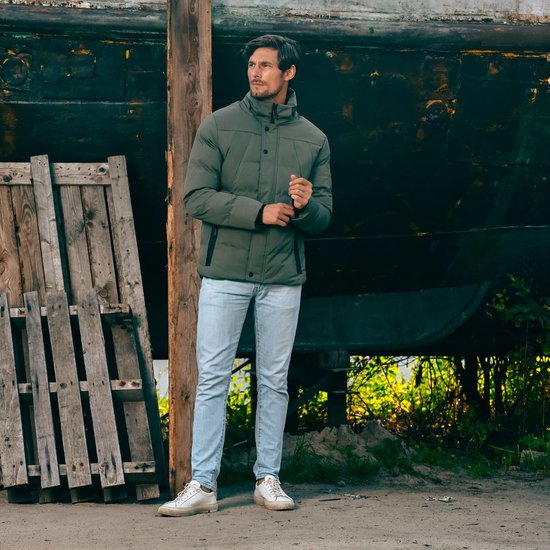 Man met olijfgroene winterjas en lichtbruine spijkerbroek, staand bij een boot.