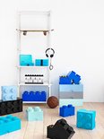 Chambre d'enfant moderne avec étagère blanche avec skateboard et casque, et diverses boîtes de rangement Lego bleues, bleu clair et noires au sol.
