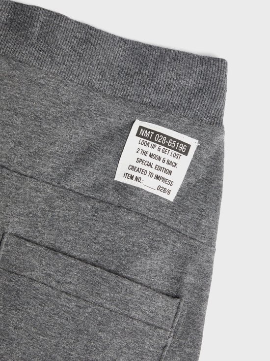 Close-up van het label op een donkergrijze joggingbroek met tekst: 'NMT 028-65196 LOOK UP & GET LOST 2 THE MOON & BACK SPECIAL EDITION CREATED TO IMPRESS ITEM NO.: 028/6'.