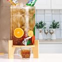 Glazen drankdispenser met houten standaard, gevuld met ijs, schijfjes citrus en donkere drank. Een glas wordt gevuld.