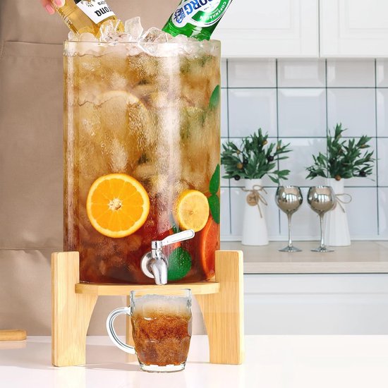 Glazen drankdispenser met houten standaard, gevuld met ijs, schijfjes citrus en donkere drank. Een glas wordt gevuld.