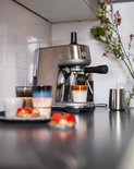 Zilverkleurige espressomachine met zwarte hendel, die koffie zet in een glas met bruine en witte banden.