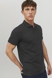 Man met donkergrijze polo en spijkerbroek, staand met één hand in de zak. Zijaanzicht.