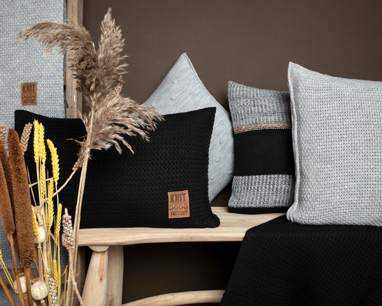 Coussins noirs tricotés avec une étiquette marron, à côté de coussins gris tricotés et de branches décoratives.