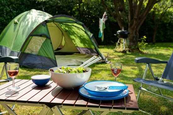 Een campingtafel met salade, borden, bestek, een glas rosé en een tent op de achtergrond.