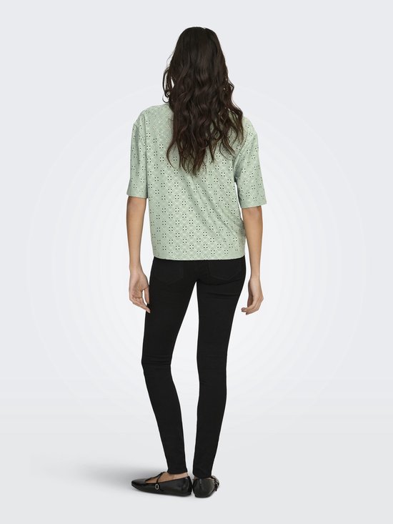 Achterkant van een vrouw in een lichtgroene blouse met broderie anglaise en driekwart mouwen, met zwarte skinny jeans.
