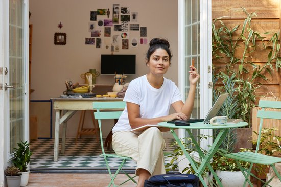 Femme en t-shirt blanc assise dehors sur une chaise verte avec un ordinateur portable, un carnet et un stylo.