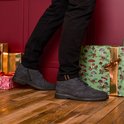 Man die donkerblauwe suède veterschoenen draagt met zwarte broek, met feestelijke cadeaus op de achtergrond.