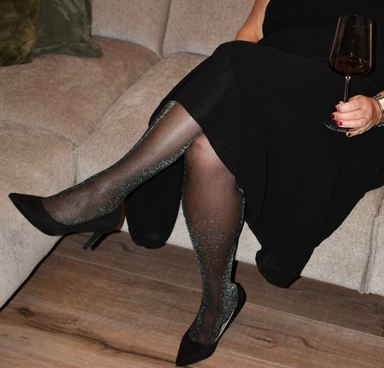 Benen van een vrouw in een zwarte jurk, met zwarte panty met zilveren glitter en zwarte pumps.